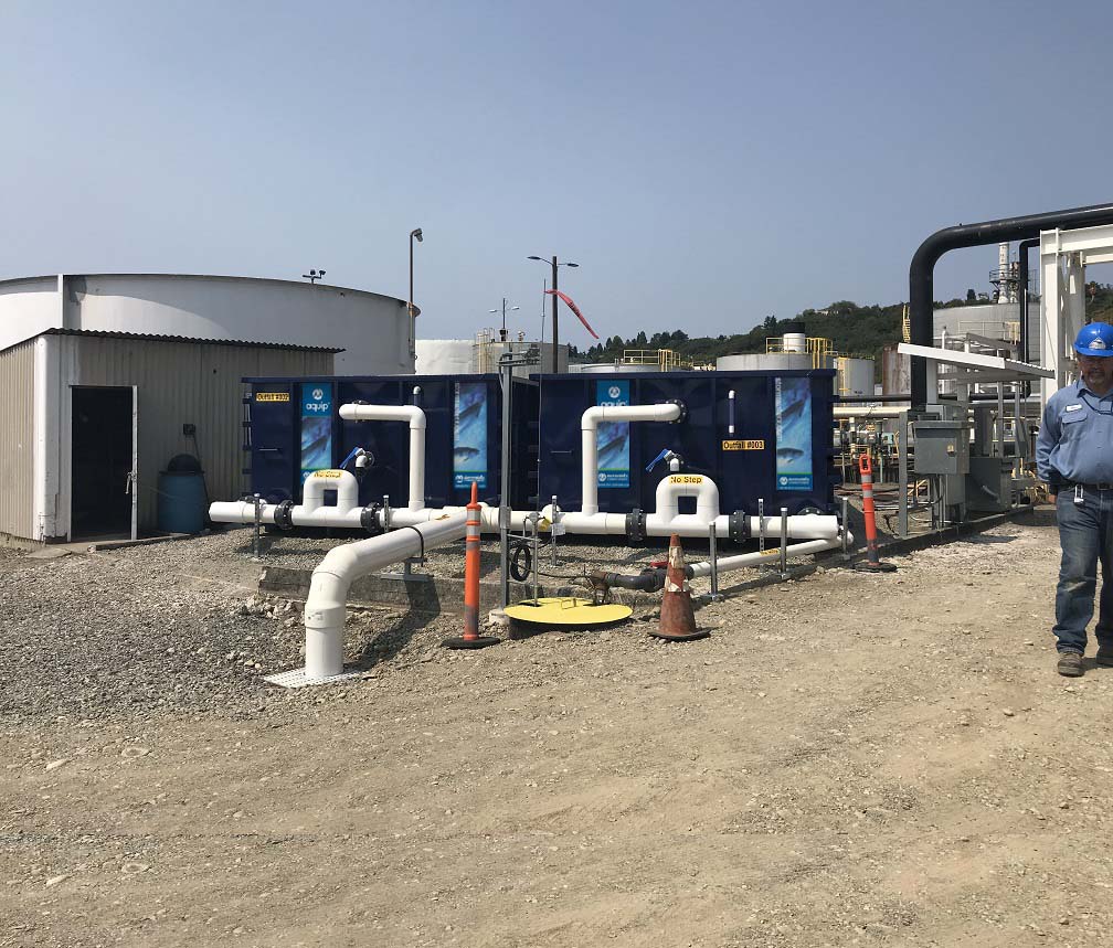 Two StormwateRx Aquip Media Filtration System in use at an industrial site consisting of two blue shipping containers connected to water sources via PVC pipes, a technician poses for the photo on the left, water tanks are in the background