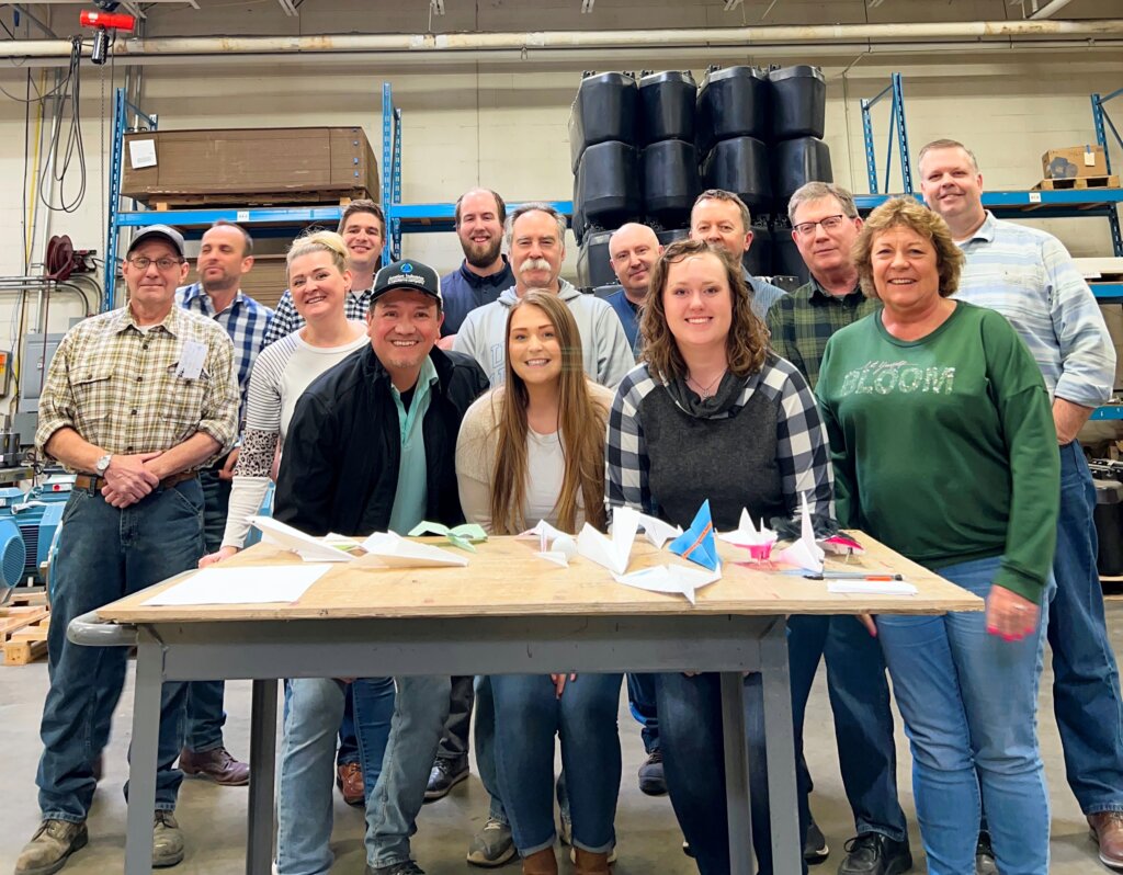 Group photo of Newterra employees in front a metal table topped with a piece of plywood covered in paper planes of various kinds, they stand in front of inventory in a warehouse, one of the women wears a green long sleeved shirt that says let yourself bloom
