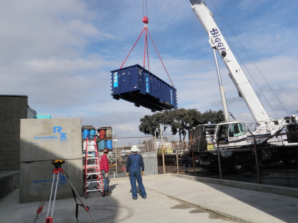 Two technician look on as a crane truck moves a Newterra Aquip filtration unit in an industrial setting, two concrete StormwateRx units are stacked behind a tripod