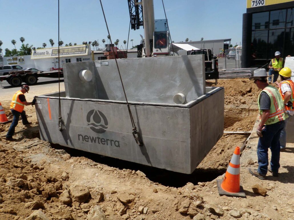 A Newterra Clara filter is lowered into a prepared dirt site at an auto recycler with four technicians guiding the unit or looking on as a crane lowers the concrete box and divider into place