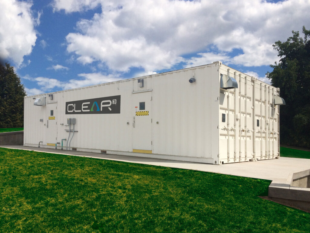 A Newterra Clear3 decentralized membrane bioreactor consisting of a white shipping container with locking doors and branded with the Clear3 logo on a concrete slab in an outdoor setting with green grass and blue skies