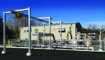 A Newterra Clear3 decentralized membrane bioreactor unit in use consisting of a beige shipping container connected to a series of chrome or steel horizontal pipes protected by a guardrail and near a frame supported hoist in a rural area.