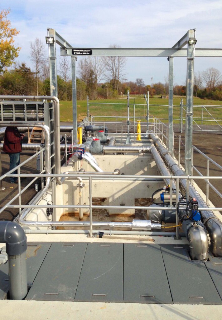 Front view of a wastewater treatment technology consisting of partitioned opened tanks connected to various kinds of pipes with a guardrail around the concrete tanks, brown treatment water is in the first tank, a technician looks at a mobile device on the left in a rural setting