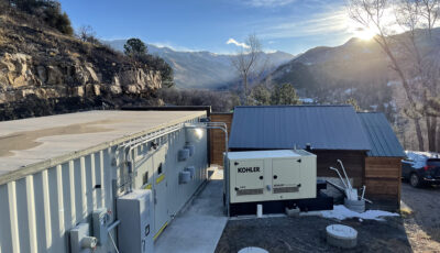 A Newterra Clear3 decentralized membrane bioreactor system in the mountains of Telluride, Colorado with the system consisting of a white shipping container with double doors, electrical panels, and meters with electrical power hooked up via a Kohler branded power unit with a small municipal building and parking area