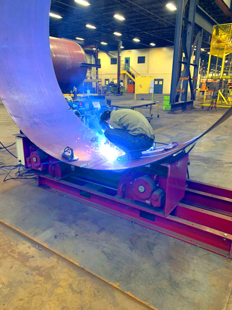 A welder creating the wall of a Newterra vessel by welding two pieces of steel together resting on a metal form, creating the initial round shape to the vessel in a production facility
