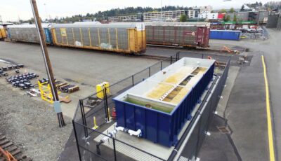 Low aerial view of a railyard with a blue Newterra modular water treatment processing unit divided into two bays connected to an underground pipe with a chain link fence around the unit, train cars sit nearby on the end of the rail line, condos and apartment buildings of a medium size city are in the background