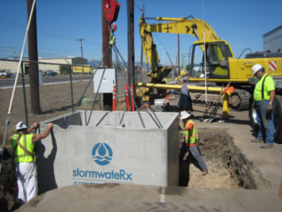 A stormwateRx unit is lowered by a crane into a sub surface opening where two technicians on opposite corners guide the open top concrete box into place, a supervisor looks on while a scoop excavator is parked in the background