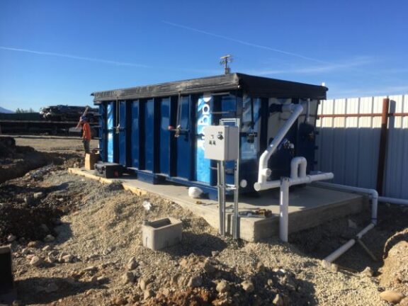 Aquip wastewater treatment unit in the field supported by a concrete slab near a fence consisting of a shipping container with various hoses extending outside of it