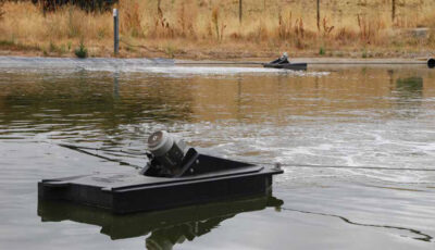 Two Newterra Aire-O2 aspirator aerator units float on an outdoor wastewater treatment pond in a rural area, the units are comprised of a flotation box with the aerator unit partially submerged with the entire float tethered to a line across the pond
