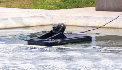 A Newterra Aire-O2 aspirator aerator floats in a wastewater treatment tank made of concrete and is held in position by steel eye hooks threaded with a heavy steel wire; green plants fill the background