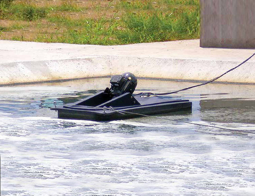 A Newterra Aire-O2 aspirator aerator floats in a wastewater treatment tank made of concrete and is held in position by steel eye hooks threaded with a heavy steel wire; green plants fill the background