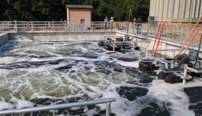 A Newterra Argos sequencing batch reactor in use with two floating aeration units mounted to metal frames in a holding tank with grated walkways and guardrails around, four wastewater engineers in the mid background view the units in operation in a rural setting, a service building and concrete water tower sit in the background