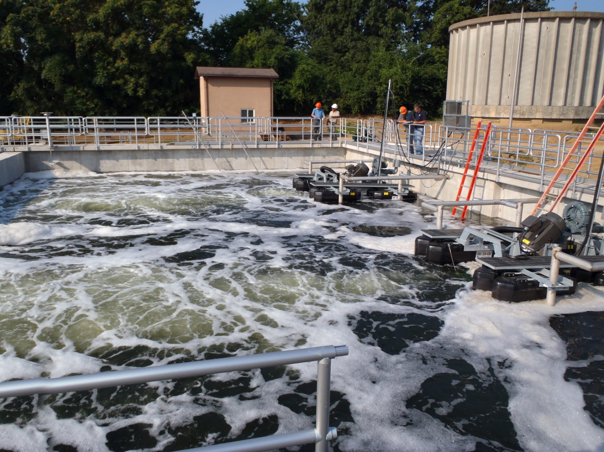 A Newterra Argos sequencing batch reactor in use with two floating aeration units mounted to metal frames in a holding tank with grated walkways and guardrails around, four wastewater engineers in the mid background view the units in operation in a rural setting, a service building and concrete water tower sit in the background