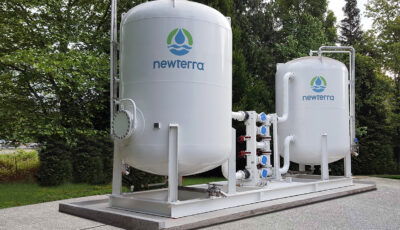 Two Cansorb high pressure liquid phase active carbon adsorbers connected by pipes filter water in a designed environment, system mounted on concrete slabs with cars in a parking lot nearby