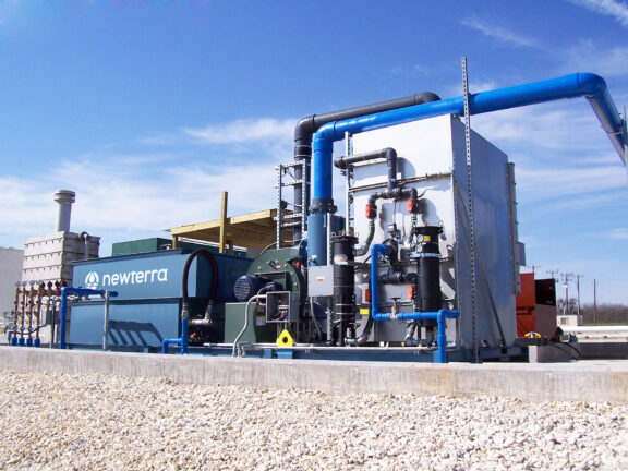Newterra vacuum extraction system consisting of a series of metal boxes connected by pipes and hoses with the center blue box branded with the Newterra logo and the box on the right connected to a blower motor, pipes of various sizes with gauges and electrical panels in an industrial setting, the unit rests on a concrete slab near power lines