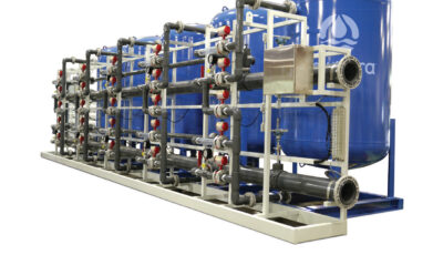 A white and blue metal frame supports a cochrane multi media filtration system consisting of five elevated blue vessels and a series of pipes in various sized containing gauges and other omponents supporting the water filtration process