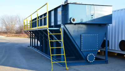 A Newterra Clara longbox filtration unit in an industrial setting consisting of a yellow ladder and guardrail attached to the unit with a V shaped base running the length of the box with a rectangular box over it, riveted access panels can be seen at the base of the unit
