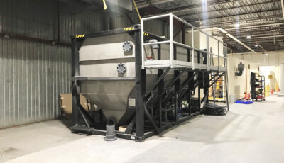 A Newterra longbox filter sits in an industrial building consisting of a metal box with a v shaped base with a black frame and a maintenance access walkway and set of stairs