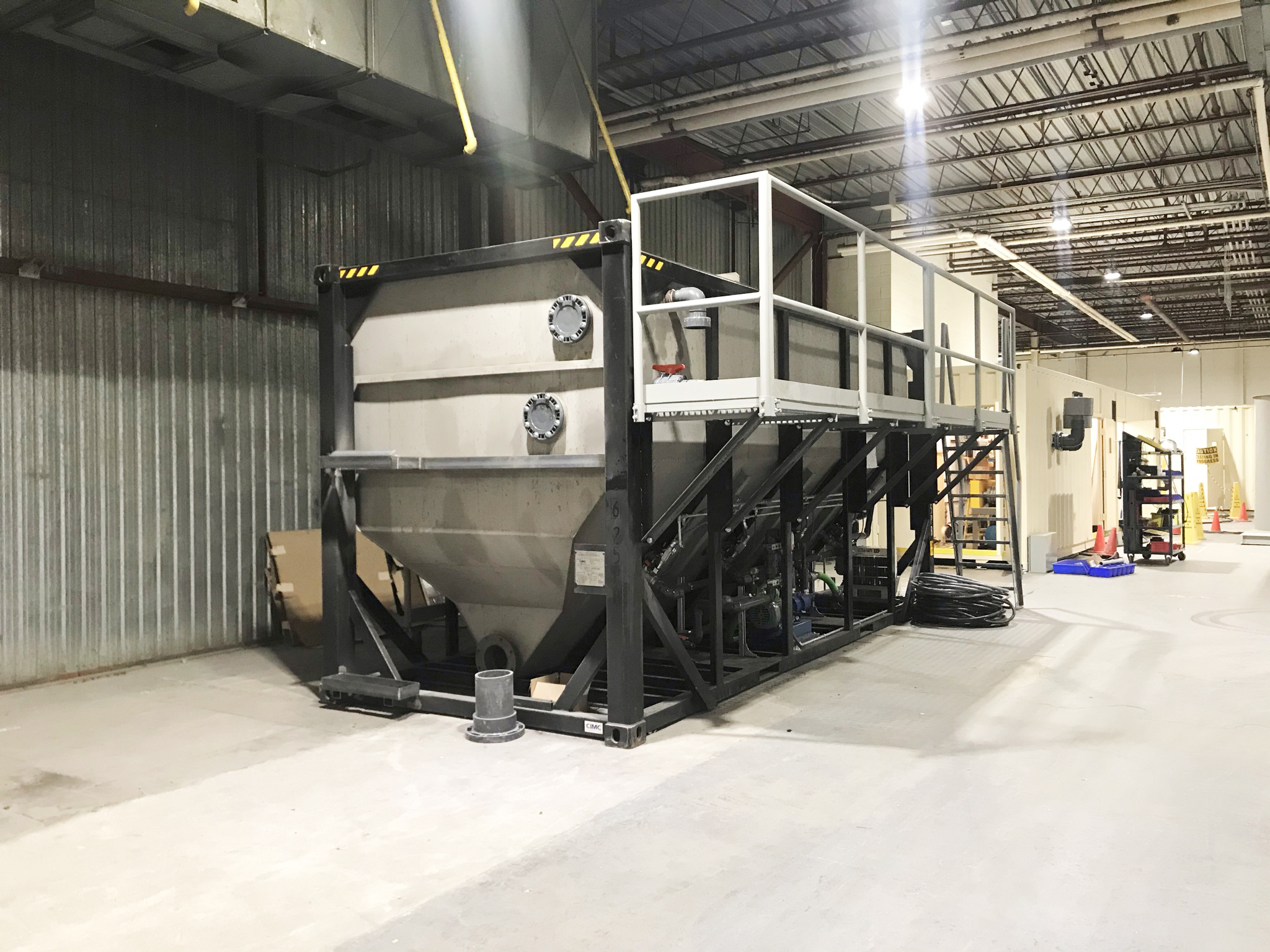 A Newterra longbox filter sits in an industrial building consisting of a metal box with a v shaped base with a black frame and a maintenance access walkway and set of stairs