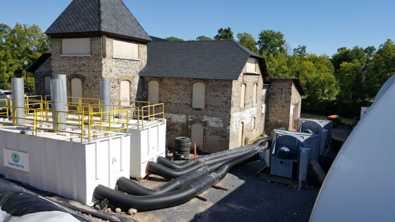Two Nixtox vapor phase box adsorbers sit next to a historic mill building with large accordion tubing entering and exiting each unit in an outdoor setting