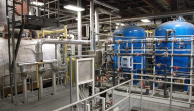 Interior view of water treatment processing equipment including a walkway for access to a horizontal metal vessel, two blue vertical vessels with equipment connected by pipes of various sizes and materials containing gauges and valves.