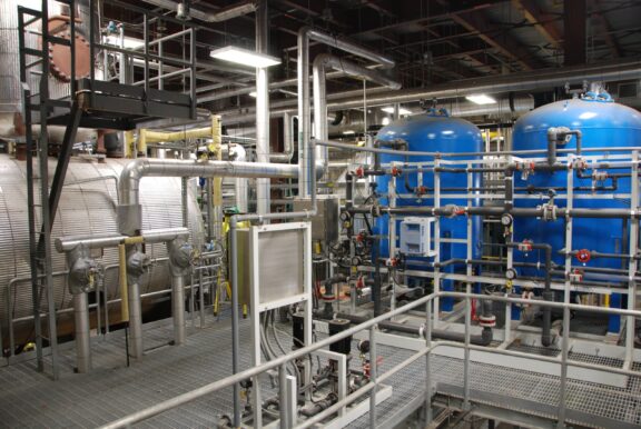 Interior view of water treatment processing equipment including a walkway for access to a horizontal metal vessel, two blue vertical vessels with equipment connected by pipes of various sizes and materials containing gauges and valves.