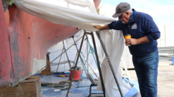 A boatyard operator pull back a white tarp to reveal the underside of a boat undergoing maintenance to demonstrate the process of how copper and zinc enter the local waterways