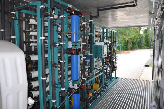 Interior view of a Seamega modular water desalination system consisiting of a cyan metal frame, various horizontally positioned filtration pipes, horizontal filters, electrical panels, and various pipes connecting the equipment to a water tank all located inside of a shipping container
