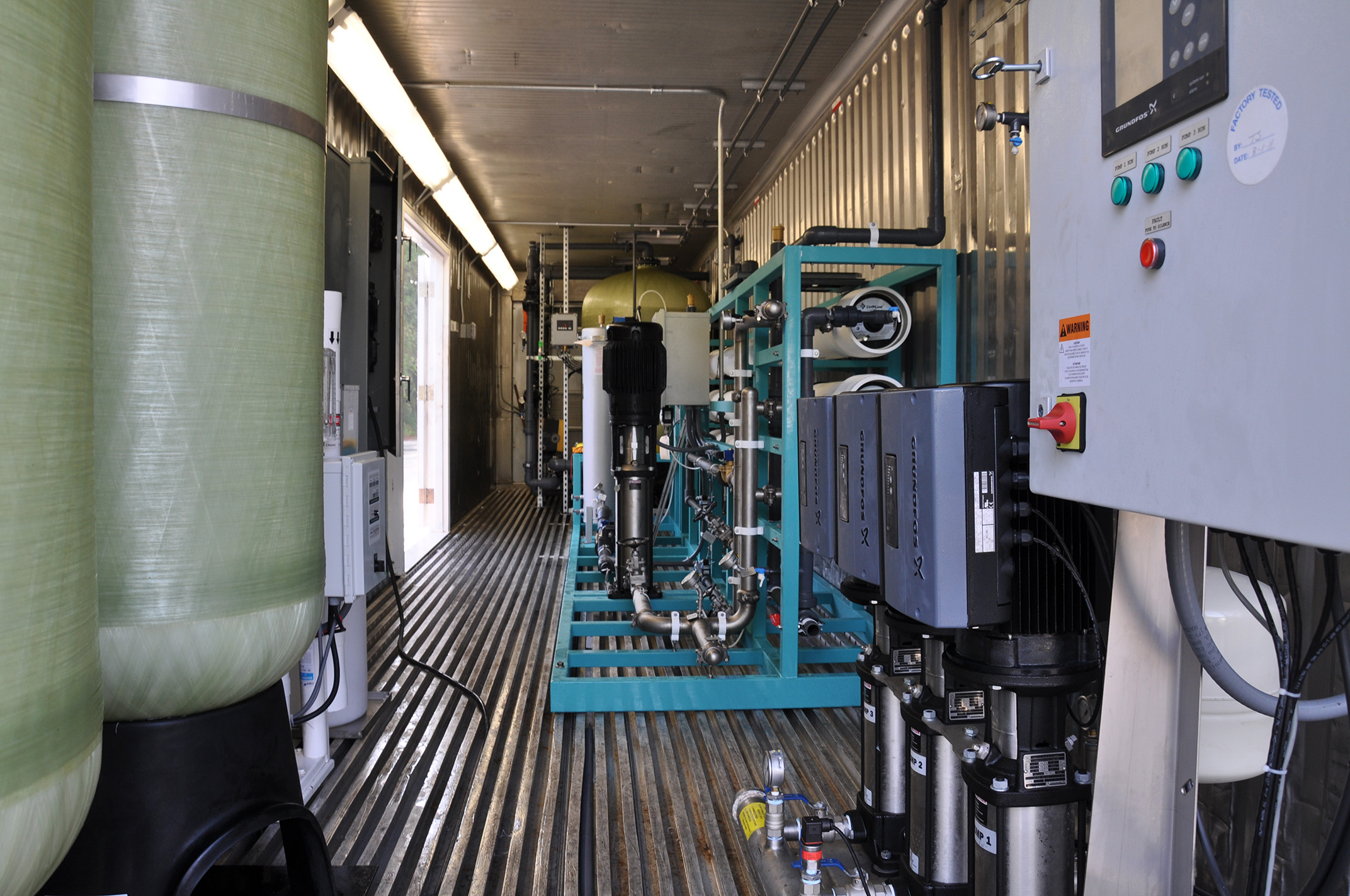 Interior view of a modular Seamega sea water desalination system including an electrical panel in the foreground across from fiberglass vessels with a cyan metal frame in the center of the image supporting a large water filter, electrical system, and various horizontally positioned filtration pipes within a metal shipping container style housing, a larger vessel stands in the back of the container