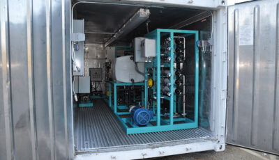 A modular mobile Seamega sea water desalination system consisting of a shipping containing housing a cyan metal frame supporting a small motor, water tank, electrical panels, and various horizontally positioned filtration pipe units, overhead lighting and additional electrical panels accompany the unit