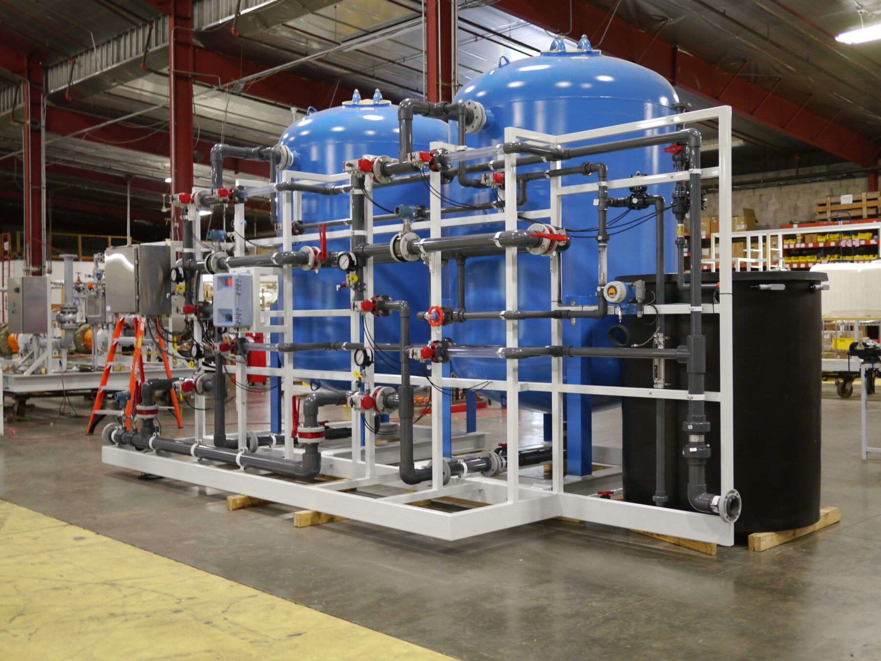 A Cochrane by Newterra water softening system sits in an industrial warehouse comprised of a black tank connected to two elevated blue vessels, the entire system connected by grey PVC pipes containing valves and other components for regulating the water softening process