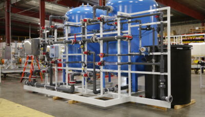 A Cochrane by Newterra water softening system sits in an industrial warehouse comprised of a black tank connected to two elevated blue vessels, the entire system connected by grey PVC pipes containing valves and other components for regulating the water softening process