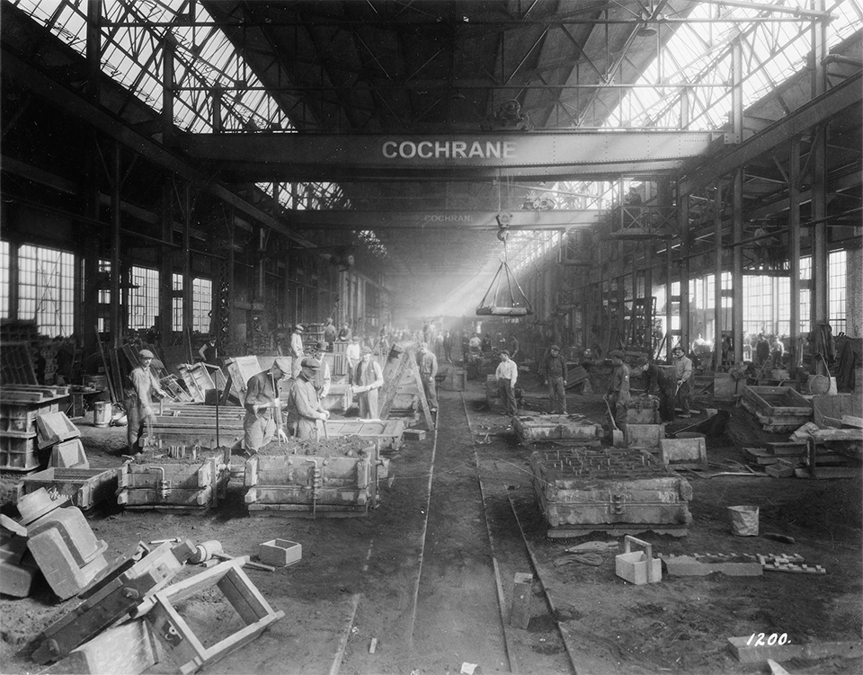 Historic image of a Cochrane dirt floor warehouse from the early 20th century prior to the installation of electricity where men are seen working with concrete molds to product Cochrane products.