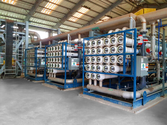Three reverse osmosis Delta units installed on a concrete slab inside an industrial warehouse with each unit consisting of horizontally oriented plastic filtration pipe units stacked within a blue metal frame along with an electrical panel and pipes of various sizes with valve and gauge components