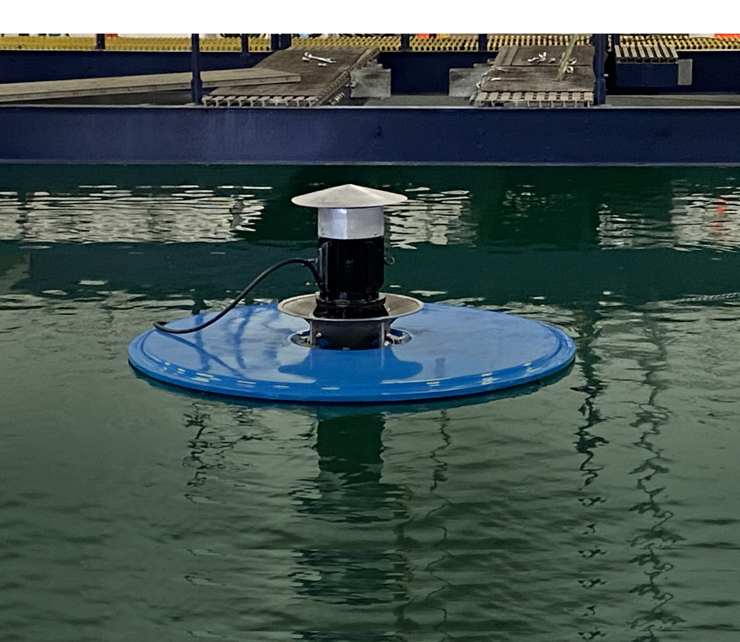 Above surface view of the Newterra Halo surface splasher aerator made of a blue metal disc with a lip and metal and black finish components terminating to a shallow metal cone as the unit aerates waste water in an indoor treatment pond