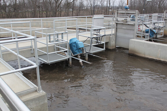 A series of concrete tanks filled with wastewater with grated metal walkways with guardrails, two aerator units are mounted to metal frames and are partially submerged and connected to power supplies via heavy flexible cables