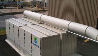 Aerial view of two Newterra air remediation modular units painted white and branded with Newterra's logo connected to a air input or output connections at the lower end and across the top of one container, the units sit next to a larger metal building ourdoors near other industrial processing equipment