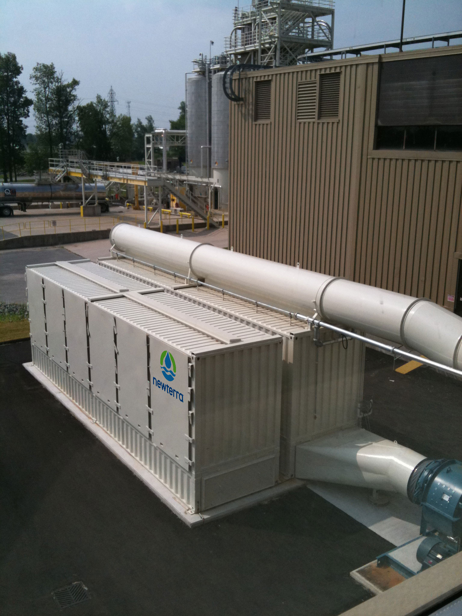 Aerial view of two Newterra air remediation modular units painted white and branded with Newterra's logo connected to a air input or output connections at the lower end and across the top of one container, the units sit next to a larger metal building ourdoors near other industrial processing equipment