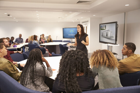 A professional woman dressed in black presents to a group of multi ethnic professionals and students in a modern presentation setting utilizing technology during the presentation