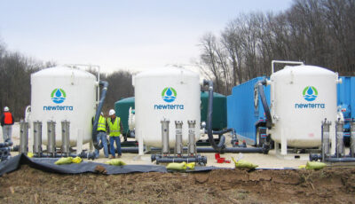 Three white Newterra branded tanks in the field process water while three technicians observe the treatment plant, an additional holding tank and modular treatment container are in the background