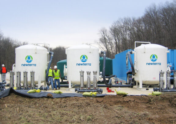 Three white Newterra branded tanks in the field process water while three technicians observe the treatment plant, an additional holding tank and modular treatment container are in the background