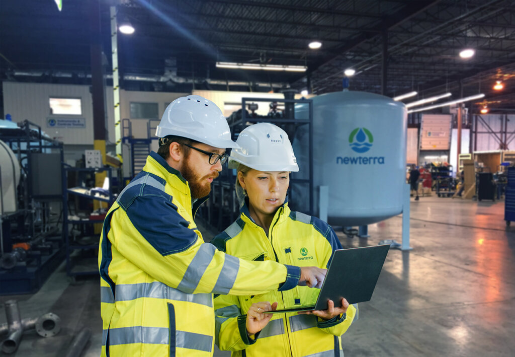 Male and female wastewater engineers consult a laptop for data in an industrial setting, a Newterra elevated tank stands in the background of the concrete floored warehouse.