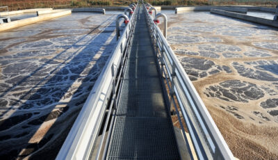Close up of a Newterra Tri Oval oxidation ditch wastewater treatment system including a grated metal walkway running the length of the system with water being treated on either side underneath the walkway, concrete divisions in the system are present on both sides of the walkway with trees in the background within a rural outdoor setting