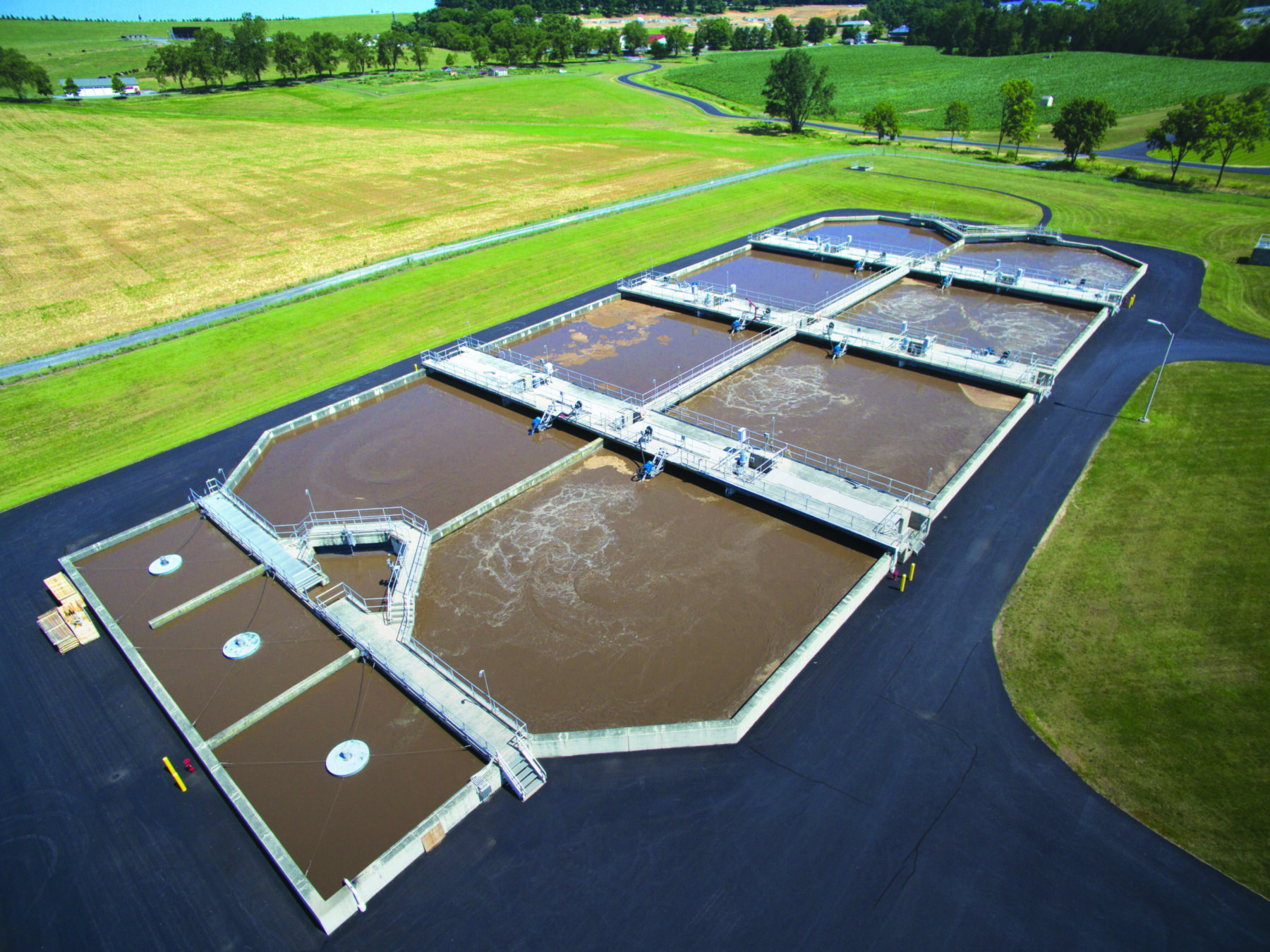 Aerial view of an open air water treatment system consisting of a series of concrete ponds connected by walkways and utilizing Newterra Trion 2.0 series process aerator mixers to stir treatment water in an open field with rural residential home and agricultural operations nearby