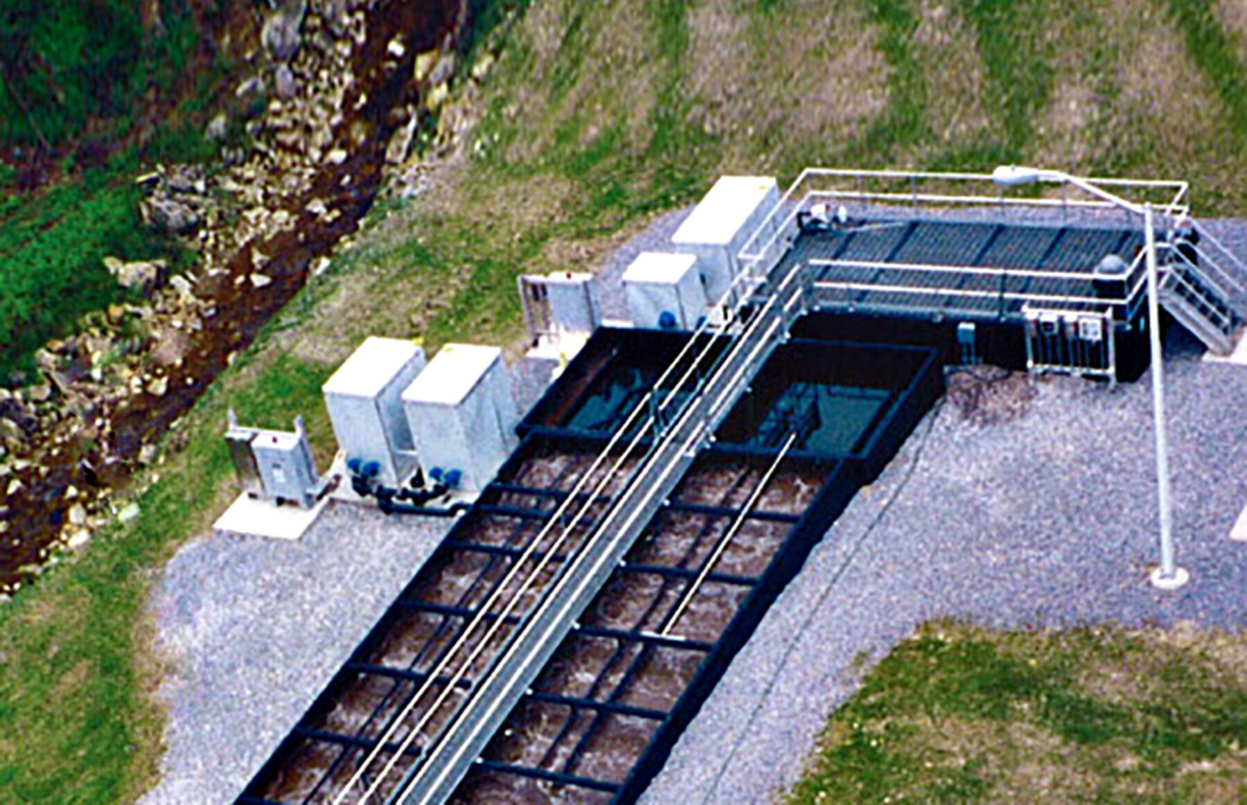 Aerial view of a Newterra Unisystem package wastewater system with a maintenance walkway across the top of the main aeration tanks and two clarification chambers, various upright metal containers and control panels are placed to the left of the treatment unit in an outdoor environment, a streetlight is on the right