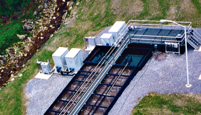 Aerial view of a Newterra Unisystem package wastewater system with a maintenance walkway across the top of the main aeration tanks and two clarification chambers, various upright metal containers and control panels are placed to the left of the treatment unit in an outdoor environment, a streetlight is on the right
