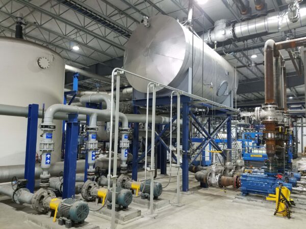 An elevated tank supported by blue steel i-beams stands above various pipes in front of the tank that connect to additional input pipes for wastewater to flow into the tank for treatment