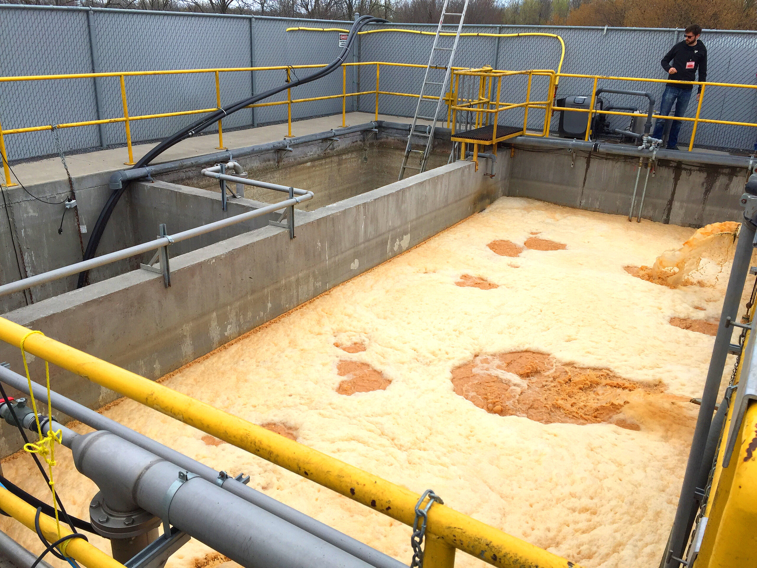 A wastewater engineer observes the wastewater treatment process of a large concrete tank with a series of divisions protected by a painted yellow steel guard rail, dirty water with tan and yellow foam enters the largest tank on the right through a pipe