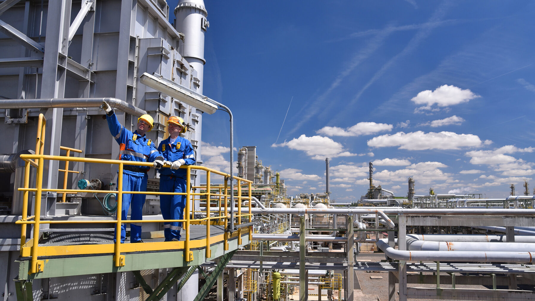 Two oil and gas workers stand on a platform overlooking the operations of the oil and gas refinery