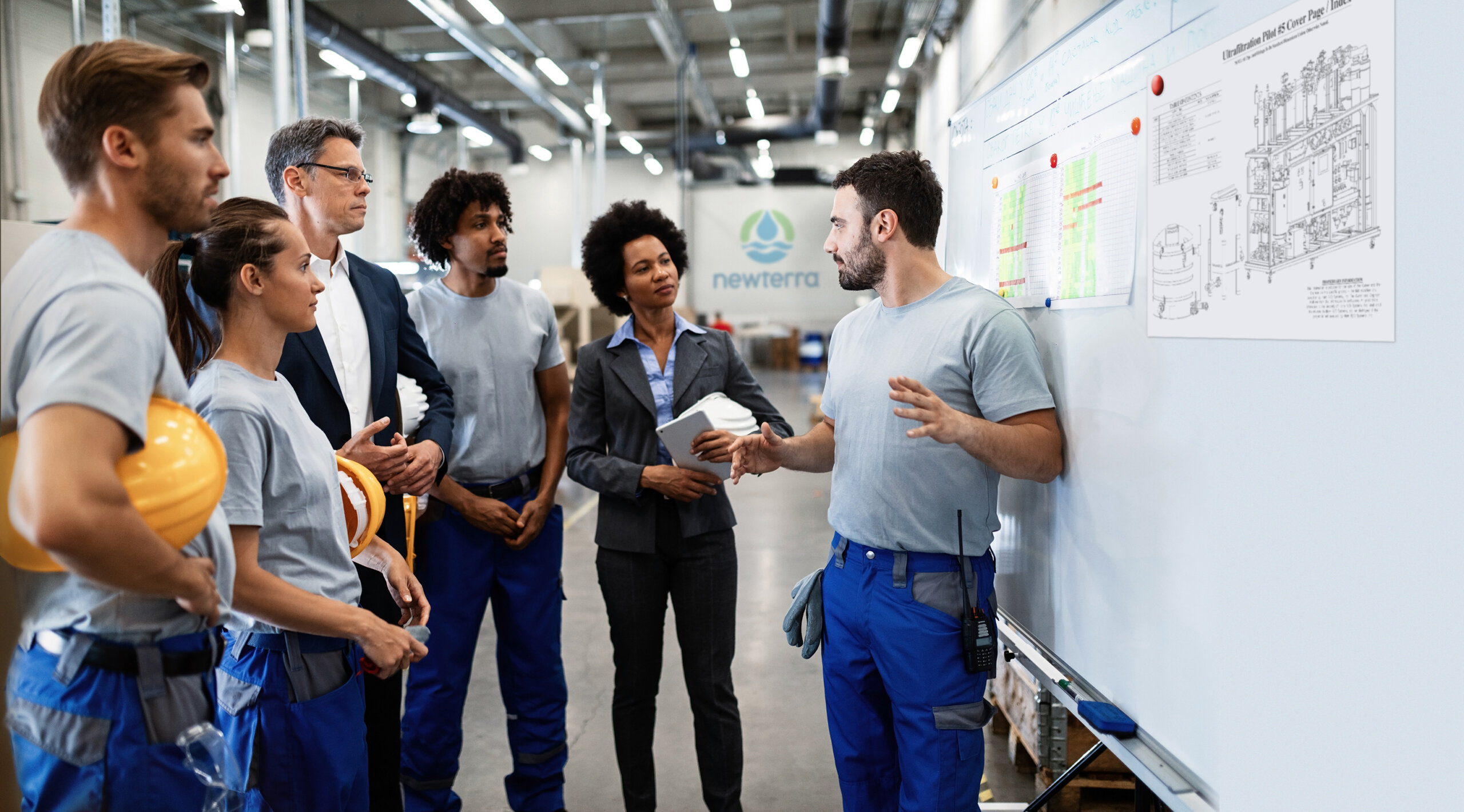 Three wastewater engineers and two wastewater professionals listen to a wastewater engineer in front of a dry erase board with equipment schematics and project notes in an industrial setting